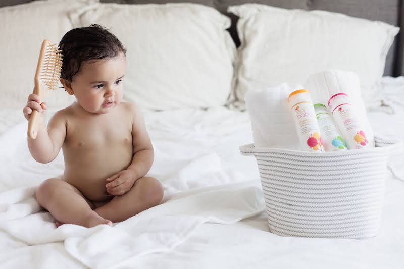 baby sitting on bed white sheets bath products holding hairbrush 