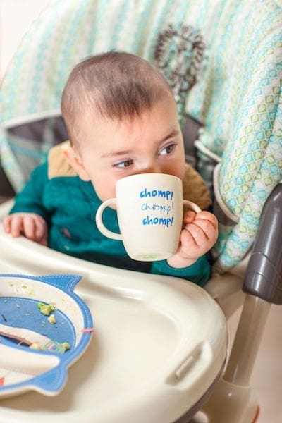 little baby drinking from white mug cup in highchair 