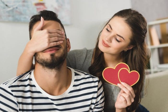 Young woman covering eyes of boyfriend with hand and holding valentine card