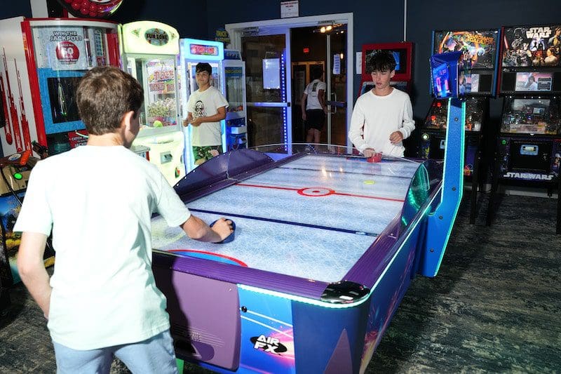 two boys wearing white shirts playing air hockey at arcade
