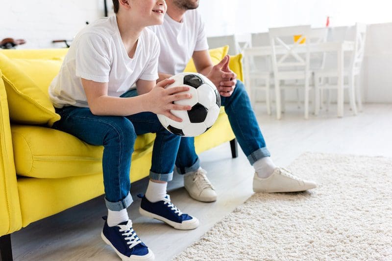 Cropped view of father and son watching sports match on couch with soccer ball