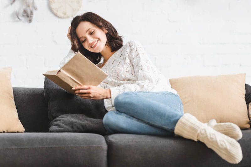 Pretty smiling girl reading book on sofa at home