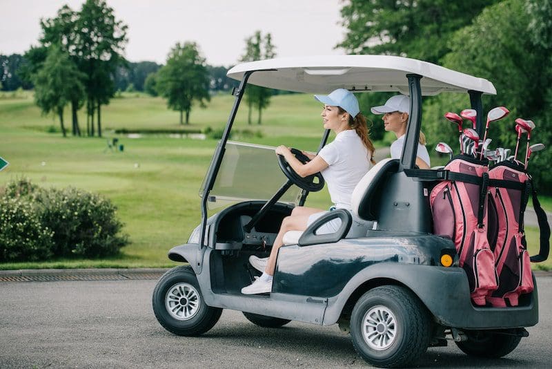 Side view of female golfers in caps riding golf cart at golf