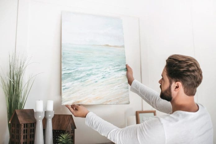 Young bearded man hanging picture on wall at home