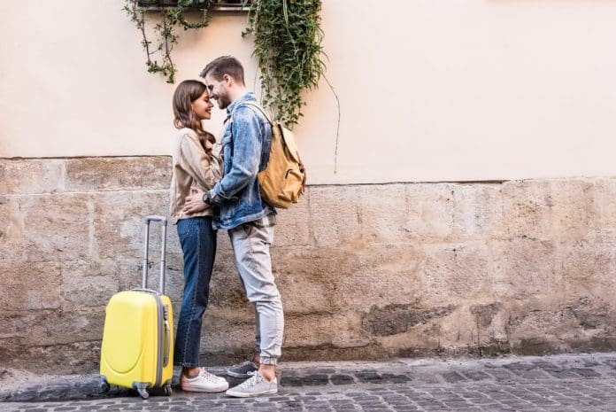 Couple with backpack and suitcase hugging near wall in city