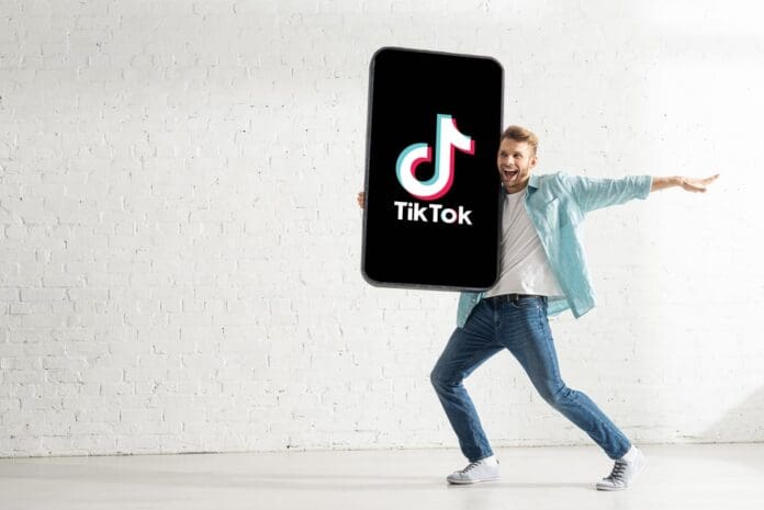 Handsome man smiling while holding big model of smartphone with TikTok app at home