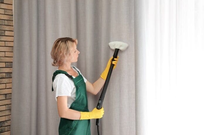 Female janitor removing dust from curtain with steam cleaner