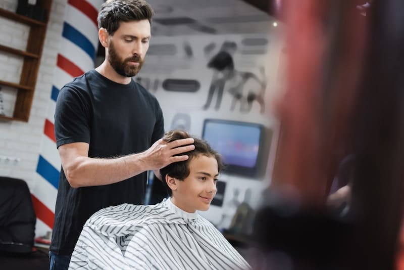 Bearded hairstylist styling hair of smiling teenage boy