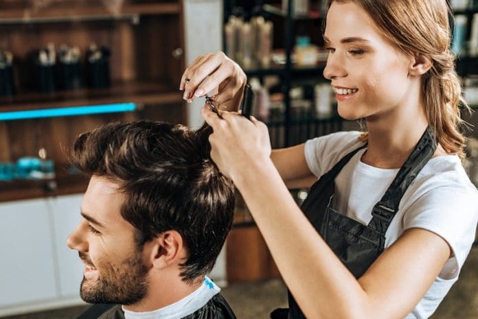 Smiling young hairdresser cutting hair to handsome happy man