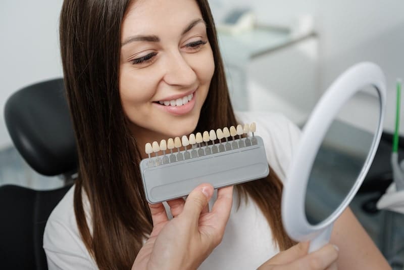 Young male dentist in white coat show veneers enamel color palette for beautiful patient woman sitting in a dental unit. Healthcare oral aesthetic treatment.